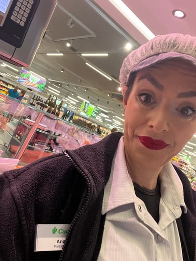 A woman with reddish lipstick, wearing a hairnet and a black jacket with a name tag reading "Cactus Ana Poissons" is standing in a supermarket. She is positioned near a scale with the words "CUCUTIED DAT BESCHT" displayed. Rows of products are visible in the background behind clear glass shelves. Overhead lights illuminate the scene. The location is likely a Cactus supermarket in Mensdorf, Luxembourg.