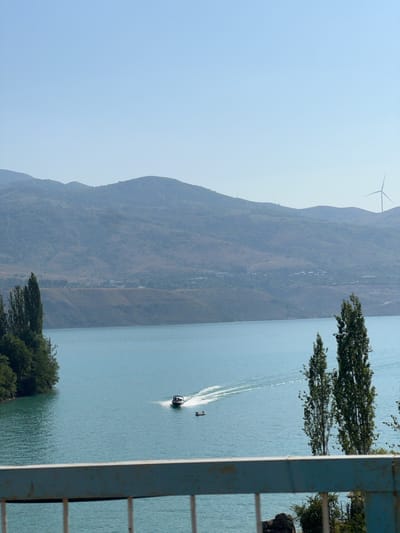 The image depicts a wide, tranquil body of turquoise water under a clear blue sky. In the distance, rolling, arid hills are visible, topped by a single wind turbine on the right side. The foreground is partially obscured by a weathered blue railing. On the water, a speed boat is creating a white wake as it moves from right to left, with two smaller figures in a raft or dinghy following behind it. The left bank of the water is lined with lush green trees. The scene is likely the Charvak reservoir, a popular recreational area near the city of Yakkatut, Uzbekistan, known for its picturesque landscapes and water activities.