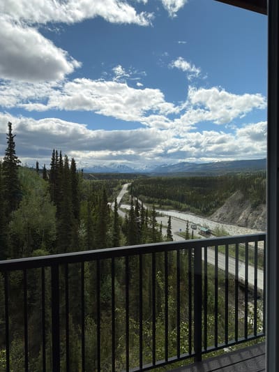 The image depicts a scenic view from a balcony overlooking a vast, forested landscape in Denali National Park and Preserve, United States. In the foreground, a black metal railing frames the vista. Beyond the railing, a dense forest of evergreen trees covers rolling hills. A winding road and a river are visible in the middle ground, with a bridge crossing the river. In the distance, snow-capped mountains are silhouetted against a sky filled with fluffy white clouds. The scene is bathed in natural light, suggesting it is daytime. There are no people visible in the image. The environment is natural and wild, characterized by trees, mountains, and a river.