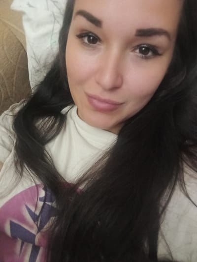 The image is a close-up, selfie-style portrait of a young woman with dark hair and makeup. She is wearing a light-colored t-shirt with a graphic design on it. The background appears to be a bed with white and patterned bedding. No specific location in the city of Yegoryevsk, Russia can be determined from this image.