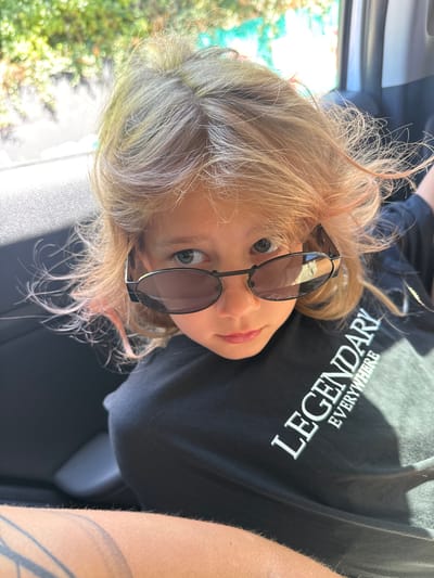A person with light blonde and gray hair, wearing dark sunglasses and a black t-shirt with the word "LEGENDARY" printed on it, is positioned inside a car. The background features greenery. The location appears to be Kruče - Kërruçi, Montenegro.