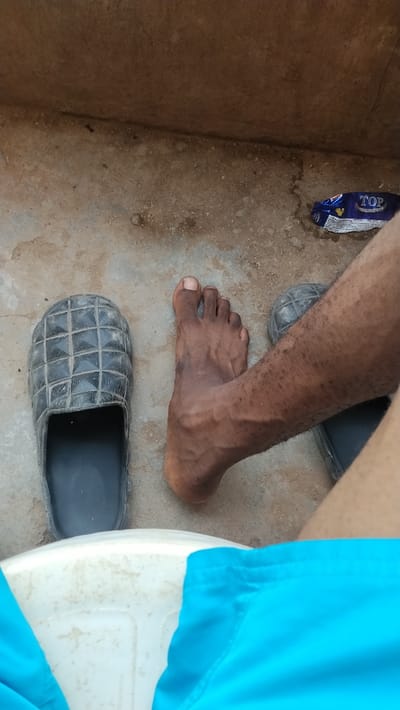 The image provides a downward, first-person perspective of a person's lower body and the immediate ground in Isieke, Nigeria. The bottom of the frame shows light blue fabric, consistent with shorts, covering the upper thighs, resting against a white, possibly plastic, surface. Ahead, on a light brown, dusty concrete or packed earth floor, is a bare, dark-skinned human right foot, exhibiting visible veins and intact toes. To the left of this bare foot, a single dark grey/black slide sandal with a raised grid pattern on its upper surface rests, appearing empty. To the right of the bare foot, a portion of a second identical dark grey/black slide sandal is visible, partially obscured by the leg. In the upper right corner, a crumpled blue and white snack wrapper, bearing the word "TOP" and a Mickey Mouse-like emblem, lies on the ground. The upper background shows a light brown concrete wall or elevated edge meeting the floor.