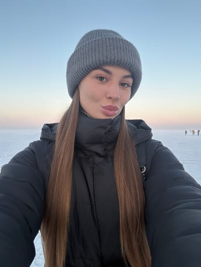 A young woman in a gray beanie and a black puffer jacket poses for a selfie in an outdoor winter setting. She has long, light brown hair and pursed lips. The background shows a pale blue and white expanse, likely snow or ice, with a subtle horizon line tinged with a warm sunset glow. Distant figures are faintly visible on the horizon. The overall impression is of a cold, serene landscape.
