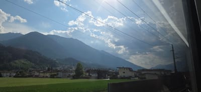 This media file captures a scenic view from a train window, likely in Inzing, Austria. The scene is dominated by majestic, hazy blue mountains that rise in the background, their peaks partially obscured by the atmosphere. In the mid-ground, a quaint village nestles at the foot of the mountains, featuring a prominent church steeple. Residential buildings with pitched roofs and various architectural styles are visible, interspersed with trees and verdant fields.

The sky above is a bright blue, dotted with dramatic, sunlit cumulus clouds that cast shadows and create a sense of depth. Sunlight streams through the clouds, creating bright shafts of light that illuminate the scene. Overhead, a network of dark power lines stretches across the frame, a common sight along railway routes, with a utility pole also visible. The time of day appears to be late morning or early afternoon, given the bright sunlight and the angle of the sun.

The overall mood is peaceful and serene, evoking the feeling of a scenic journey through the Austrian countryside. The lush green fields in the foreground suggest a time of year when vegetation is in full bloom, likely late spring or summer. There is no visible text in the image.