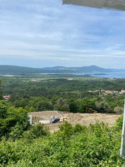 The image depicts a view from an elevated position in Kavač, Montenegro. In the distance, mountains and a body of water are visible under a sky with scattered clouds. In the middle ground, dense green vegetation covers the land. A dirt construction site is present, featuring an excavator and some workers. A concrete structure is also visible in the construction area. Green foliage is prominent in the foreground.