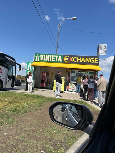 The image shows a street scene in what is presumed to be Giurgiu, Romania, featuring "La Vinieta Exchange Rovinieta". A group of approximately 10 people are lined up outside the exchange booth. A street light stands on the left side of the exchange. A bus with the marking "OTur" is partially visible in the foreground. A car side mirror is visible in the upper right corner, indicating the photo was taken from inside a vehicle. On the upper left of the frame, is another street sign in Romanian. An Illy coffee vending machine is visible next to the booth.