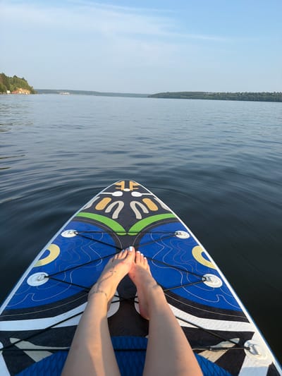 The image depicts a point-of-view shot from a paddleboard on a body of water, likely the Sylva River in Sylva, Russia, given the surrounding landscape. The foreground is dominated by the nose of a paddleboard featuring a vibrant blue, black, and white design with green and yellow accents. The camera angle is looking forward, with the paddleboard's textured deck clearly visible. Two bare feet, with light blue painted toenails and a delicate ankle tattoo of interconnected circles, rest on the paddleboard. The water is calm, with gentle ripples reflecting the pale blue sky. In the mid-ground, distant tree-covered shores form a natural horizon line, with a hint of buildings visible on the left bank. The overall scene evokes a sense of peaceful recreation on a serene natural waterway.