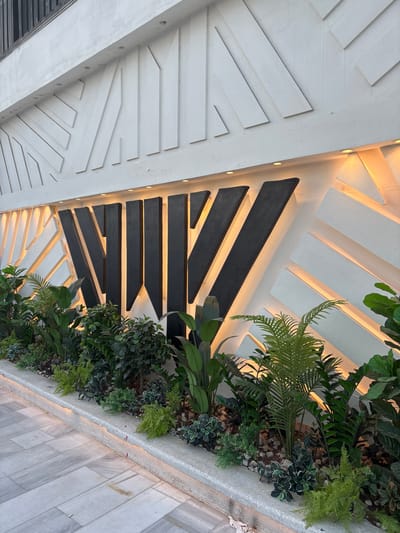 The image shows a modern building facade in Khirbet Al Souk, Jordan. The facade features a white textured wall with geometric, angular relief patterns. Across the upper portion of the wall, there are dark, angled, three-dimensional letters that appear to spell "WAV". These letters are illuminated from behind with warm yellow light, creating a dramatic effect. Below the wall is a manicured garden bed filled with various green plants, including ferns and broad-leafed foliage. The garden bed is bordered by a concrete planter box which is subtly lit from underneath with the same warm yellow light, casting a glow on the adjacent walkway. The walkway is paved with large, light-grey rectangular tiles laid in a linear pattern. The scene appears to be outdoors, possibly in the evening or early morning, given the artificial lighting.