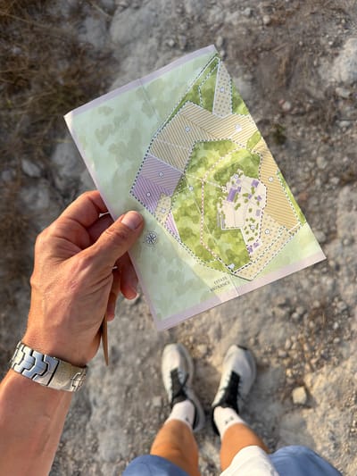 A person holds a map of an estate entrance at ground level. The map is unfolded and displays a stylized representation of a property with numbered sections, green areas, and buildings. The person's hand, wearing a silver watch, is visible on the left side of the frame. Below, the legs and feet of the person are visible, wearing white socks and athletic shoes. The ground is covered in gravel and dry vegetation. The scene appears to be outdoors, in a rural or natural environment. The location is identified as Montuïri, Spain, suggesting the estate is in this region.