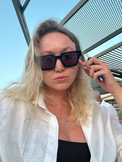A woman with blonde hair and sunglasses is taking a selfie outdoors. She is wearing a white unbuttoned shirt over a black top and a necklace. She is holding a purple electronic cigarette device to her ear with her right hand. The background shows a blue sky and a metal structure with a lattice pattern. The scene appears to be in Plataniás, Greece, suggesting a warm, sunny climate.