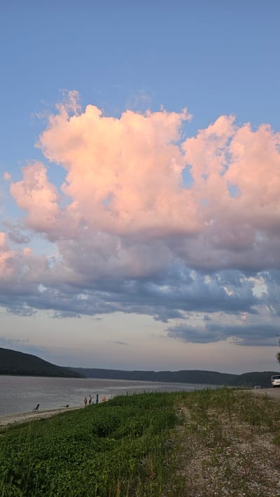 The image depicts a serene riverside landscape in Kulishivka, Ukraine, at dusk. A wide river dominates the left side of the frame, with a lush green embankment extending along its bank. A few people are visible on the sandy shore, some appearing to be fishing. The background features rolling hills covered in dense forest, receding into the distance. The sky above is a canvas of soft blues and grays, with dramatic clouds tinged with the warm hues of the setting sun. A dirt path runs along the right side of the embankment, leading towards a white van parked in the distance. The overall atmosphere is tranquil, capturing the beauty of nature as day transitions into night.