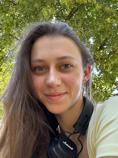A young woman is captured in a close-up selfie from a low angle outdoors, with lush green foliage of trees filling the background, suggesting a park or natural setting in Erlenbach am Main, Germany. She has long, light brown hair that falls to her left side, framing her face. Her blue-green eyes are looking directly at the camera. She is smiling gently, with her lips slightly parted. She wears a light yellow top, and a black pair of Marshall headphones are resting around her neck. A delicate gold earring with a star-like charm is visible on her right ear. The lighting suggests it is daytime, with sunlight dappling through the leaves, creating a bright and natural atmosphere. The scene is devoid of any other people or specific landmarks that would pinpoint a more exact location within the city.