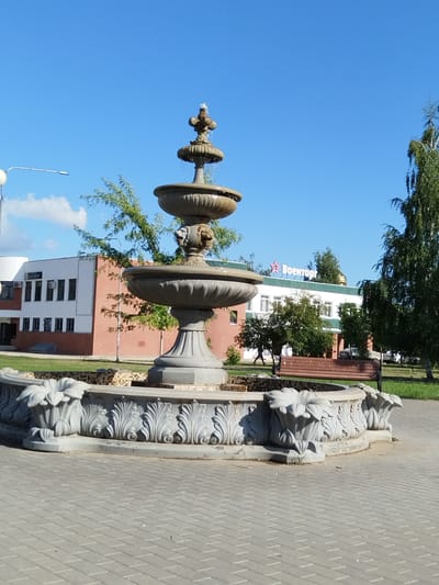 The image shows a multi-tiered stone fountain in the foreground, with intricate carvings around its base. The fountain is located on a paved area, possibly a plaza or park. In the background, there are buildings, trees, and a clear blue sky. One building has a red star and Cyrillic text that reads "Военторг" (Voentorg), indicating it is likely a military trade store. A wooden bench is visible to the right of the fountain. There are no people clearly depicted in the scene, though a faint silhouette of a person walking can be seen in the mid-ground. The environment appears to be an outdoor urban or suburban setting on a sunny day. The location is identified as Roshchinsky, Russia.