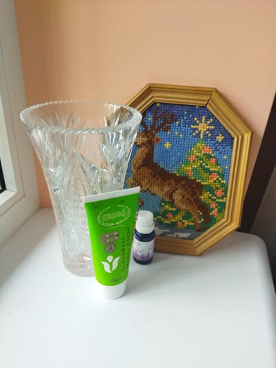 A still life scene is depicted on a white surface against a peach-colored wall. To the left, a tall, clear glass vase with intricate cut patterns is positioned. In front of the vase, a green tube of "ERSAG CLEANBALL MASAJ KREMI" is placed, and to its right, a small dark purple bottle with a white cap is situated. Behind these objects, a framed cross-stitch artwork featuring a reindeer and a Christmas tree under a starry sky is leaning against the wall. The artwork has a hexagonal wooden frame. The lighting suggests an indoor setting. There are no people or animals visible in the image. The location in Dolyna, Ukraine, is not discernable from the visual information provided in the image.