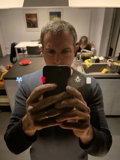 The image depicts a mirror selfie taken by a male individual in an indoor residence located in Foetz, Luxembourg. The man, appearing in his 30s or 40s with short greying hair, wears a dark blue long-sleeved top. He holds a black smartphone with both hands, positioned to capture his reflection. A plain gold band is visible on the ring finger of his left hand.
Reflected in the background is a domestic kitchen and dining area. To the right of the man's reflection, a dark-haired individual is partially visible behind a dark kitchen countertop, appearing to be focused on a device or object. The counter displays a sink, faucet, yellow cloths, and a pot containing green produce. To the left, a white dining table with dark chairs, a red circular object, and a bowl of apples are visible. Two framed artworks hang on a light-colored wall behind the dining area.
Several luminous, abstract shapes are present on the reflective surface, including a blue star-like symbol on the left side of the man's chest, a red segmented rectangle near his right shoulder, a white gear-like symbol, and a small white rectangle further to the right. The overall lighting suggests an interior, possibly evening, setting.