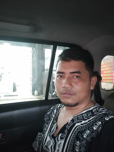 A mid-adult man with short, dark hair and a short beard is seated in the rear passenger compartment of a vehicle in the city of Namplohbaro, Indonesia. He is wearing a black collared shirt adorned with a repeating white geometric and organic pattern, and looks directly at the camera with a neutral expression. The vehicle's interior features dark door panels and a light gray headliner, with a beige front passenger seat partially visible to the right. Through the left rear window, a brightly lit outdoor scene reveals a building with a glass door or large window, a partial view of a person inside, and a white plastic jug on the ground. Through the right rear window, an exterior sign with inverted Indonesian text is visible, indicating a printing shop that offers "PERCETIKAN" (Printing), "SABLON" (Screen Printing), "CILID BUKU" (Book Binding), "COVER BUKU" (Book Cover), "UNDANGAN" (Invitations), "NEON BOX", and "PAMFLET" (Pamphlet). The man appears to be posing for a photograph.