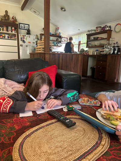 In Thora, Australia, this image captures an indoor domestic scene. A young girl is seated on a black leather couch, engrossed in drawing in a notebook. She is wearing a gray sweatshirt with red and yellow lettering on the sleeve that reads "GHOSTS". She holds a white pen in her right hand, with her left hand resting on the notebook. The notebook is open to a page with a blue drawing. In front of her on a patterned tablecloth are a remote control, a small pack of cigarettes, a rectangular pastry on a colorful tray, and a white plate with a sandwich and sliced avocado. To the right of the girl, another person's hands are visible, also interacting with the food on the plate. The background reveals a kitchen area with wooden cabinets, a refrigerator, shelves stocked with kitchenware, and a man in a dark blue tank top standing at a counter looking out a window. Sunlight streams in from the window, illuminating the kitchen. The overall environment suggests a casual family gathering or a quiet afternoon at home.