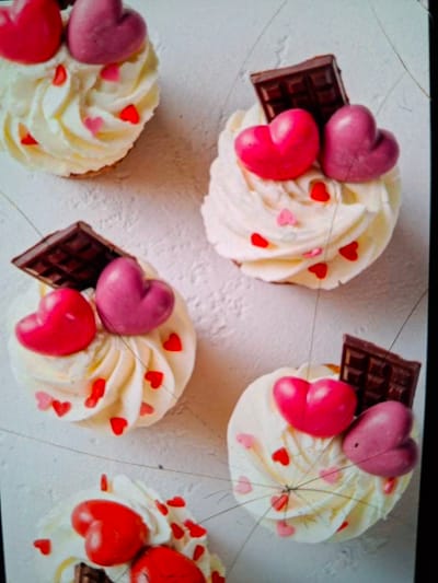 This is a close-up, overhead shot of several cupcakes on a white, textured surface, likely a table. The cupcakes are decorated with swirls of white frosting, red and pink heart-shaped sprinkles, and some are topped with chocolate pieces and larger, heart-shaped candies. The heart candies appear to be a light pink or purple and a bright pink or red. The chocolate pieces are dark brown, segmented pieces. The scene is well-lit, and the focus is sharp on the cupcakes. There is no visible text in the image. The setting suggests a celebratory occasion, possibly Valentine's Day, given the heart motifs. The overall mood is festive and sweet. The image appears to be taken indoors.