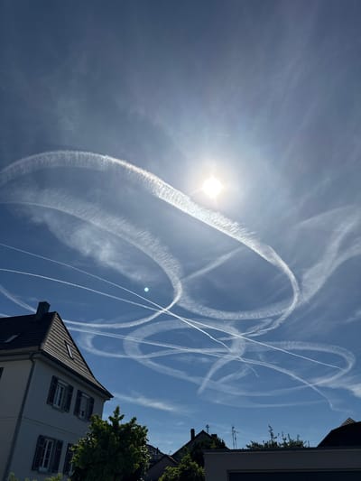 The image shows a blue sky with numerous airplane contrails forming intricate patterns, including loops and straight lines. The sun is visible, slightly obscured by thin clouds, in the upper right portion of the frame. In the lower left corner, the roof and upper portion of a light-colored building with dark shutters are visible. A small green tree is positioned in front of the building. The scene appears to be an outdoor view from a residential area in Salach, Germany. There are no people or animals visible in the frame. The overall impression is one of a clear day with significant air traffic.