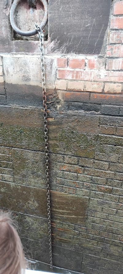 In the city of Sapotskinski Sel'ski Sovet, Belarus, a close-up, vertical shot reveals a thick, weathered metal chain suspended from a large, round metal ring affixed to a stone structure. The chain hangs down a brick wall that is partially covered in a dark, grimy substance, likely algae or moss, indicating exposure to moisture. The brickwork itself is varied in color, ranging from reddish-brown to lighter, weathered shades, with some areas showing damage and chipping. At the bottom left corner of the image, the edge of a person's head with light brown hair is visible, suggesting they are looking down at the chain or the wall. The overall impression is one of age and exposure to the elements, possibly in a waterfront or canal setting.