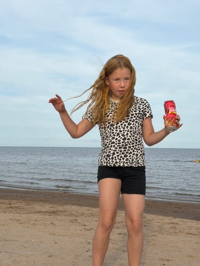 A young girl with blonde hair is standing on a sandy beach, holding a red can of what appears to be a beverage. She is wearing a black and white leopard print t-shirt and black shorts. The can has Russian text on it, including "КВАС". The background consists of a calm ocean with gentle waves and a pale blue sky. The lighting suggests it is daytime, likely late afternoon or early evening given the soft light. The girl's expression is somewhat neutral, perhaps a bit pensive or curious. The overall scene is one of leisure and relaxation by the sea. The image is oriented vertically, with the girl positioned on the left side and the sea and sand stretching out to the right. The image seems to be taken in Ondvor, Russia.
