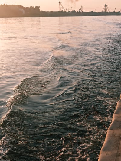 This is a photograph taken from a boat moving on a body of water, likely a river, during sunset or sunrise. The water is dark and choppy, with visible wake and ripples. The sky is a soft orange and pink hue, reflecting on the water. In the distance, a silhouetted shoreline is visible, featuring trees and industrial structures such as cranes and smokestacks, suggesting an industrial or port area. A portion of the boat's railing or deck is visible in the lower right corner of the image. The location is identified as Lesosibirsk, Russia, which is known for its timber industry and is situated on the Angara River.