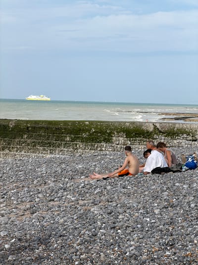 A group of people are gathered on a pebbly beach on a sunny day. In the foreground, a young boy in orange and black swim trunks bends over. Behind him, an older man in a white shirt is also bent over, with a woman in a grey top and blue bikini top leaning in towards him. Another woman, mostly obscured, is to their left. A blue backpack and other items are scattered near them on the pebbles.

The scene overlooks a body of water, likely the sea, with waves breaking against a stone and moss-covered barrier. In the distance, a yellow and white ferry is visible on the water. The sky is a clear, pale blue with a few wispy clouds. The overall impression is of a casual, relaxed day at the beach, possibly late morning or early afternoon given the bright light. The location is identified as Hautot-sur-Mer, France. No visible text is present.