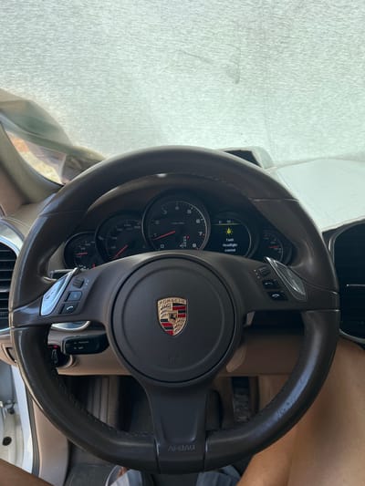 This media file shows a close-up view of the interior of a Porsche, focusing on the steering wheel and dashboard. The steering wheel is dark brown leather with silver accents and the Porsche emblem is prominently displayed in the center. Various controls are visible on the steering wheel, including buttons for media and phone functions, as well as a cruise control stalk.

The dashboard features a set of analog gauges, including a speedometer and tachometer. A digital display within the instrument cluster shows a "Fault Headlight control" message, indicating a potential issue with the vehicle's lighting system. The sun appears to be shining brightly, casting shadows on the dashboard and suggesting it is daytime.

The background reveals hints of the car's interior, with tan leather seats and a dark-colored dashboard. Part of a person's arm and leg is visible on the right side of the frame, suggesting the driver or a passenger is present. The overall impression is that of a modern luxury vehicle, with a focus on the driver's interface. The context provided indicates the location is Voysil, Bulgaria.
