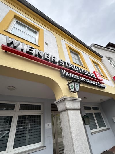 The image is a low-angle shot of a building in Amstetten, Austria. The building has a yellow and white facade, and windows with yellow frames. A sign displaying "WIENER STÄDTISCHE VIENNA INSURANCE GROUP" is vertically mounted on the building's corner. A dual-sided lit display with "Gösser" logo is attached to the sign's mounting. To the right is a gray, modern building segment with white-framed windows, and Venetian blinds are visible. The sky is overcast.