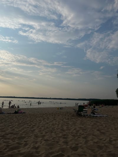 A wide shot of a sandy beach on a cloudy day. Numerous people are in the water, some swimming and others wading. On the shore, people are relaxing on towels, and there are a couple of beach chairs. The water extends to a distant tree line under a sky with scattered clouds and sunbeams.