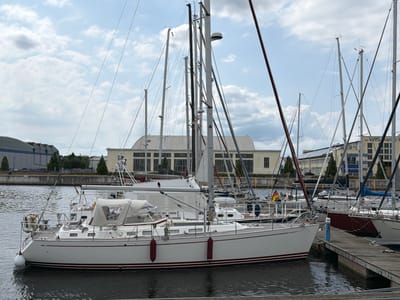 The image shows a marina in Wismar, Germany, with several sailboats docked side-by-side. The foreground features a white sailboat with a red stripe along its waterline, equipped with a Bimini top and various deck fittings. The boat is moored to a wooden dock. In the background, more sailboats with tall masts are visible, along with buildings that appear to be part of a harbor infrastructure. The sky is cloudy, suggesting overcast weather. There are no people clearly visible in the scene. The overall impression is of a quiet, functional harbor.