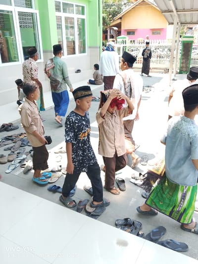 An outdoor scene captured in Jagapura Kulon, Indonesia, depicts approximately 10-15 individuals, primarily males ranging from young boys to adults, congregated on a bright, sunny day at 12:25 PM. The scene is set adjacent to a light green building featuring multiple white-framed windows and white steps, likely a mosque or prayer hall.
In the foreground and midground, multiple individuals are walking. A young boy in a black-and-white patterned t-shirt, black pants, and a black peci with gold trim walks barefoot. To his right, another boy in a light brown shirt and dark brown pants adjusts a red-fringed head covering with both hands, also barefoot. A third boy, to the left of the central pair, wears a light brown shirt and dark brown pants, standing on blue flip-flops and holding a black object. Throughout the paved area, approximately 50-70 pairs of sandals and flip-flops of various colors are scattered on the ground, suggesting individuals have removed them before entering a specific area.
Adult males are visible behind the boys, dressed in traditional attire including patterned sarongs, long-sleeved shirts, and peci. One adult wears a light green patterned shirt and blue sarong; another wears a white shirt, patterned sarong, and a patterned cloth draped over his shoulder. In the background, a person sits on the steps of the green building. Further back, a white and green painted metal fence separates the immediate area from distant residential buildings, one appearing yellowish-orange and another pink. The ground transitions from clean white tiles in the immediate foreground to a grey paved surface where the footwear is accumulated. Strong shadows indicate bright midday sunlight.