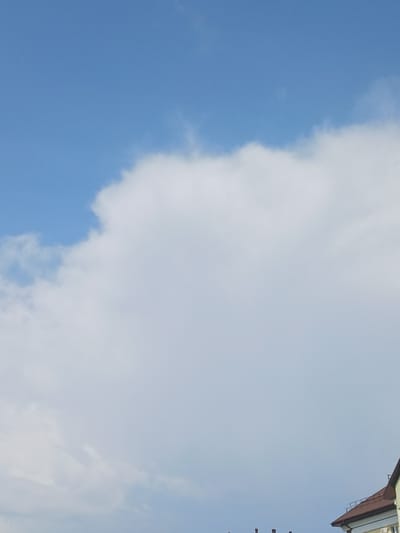 The image shows a bright blue sky with large, fluffy white clouds dominating the upper half. The lower portion of the image reveals the rooftops of several buildings, characteristic of urban architecture. One building on the right has a brown tiled roof and a light-colored facade. There are also several chimneys visible on other rooftops. The scene appears to be a daytime outdoor view, with a clear sky suggesting fair weather. No people or specific activities are depicted. The location is identified as Elitnyi, Russia.