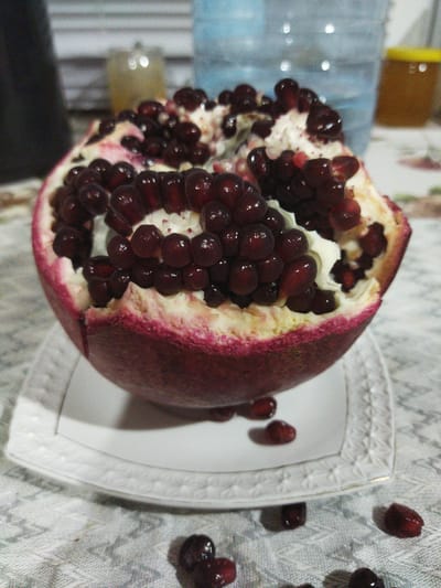 A half-cut pomegranate sits on a white decorative plate, which is on a patterned tablecloth. The pomegranate is rich with dark red arils. Several arils have fallen onto the tablecloth around the plate. In the background, out of focus, are a large blue water jug and a small jar with a yellow lid, possibly containing honey. The scene appears to be indoors, likely in a domestic setting, possibly in the city of Кіраўскі сельскі Савет, Belarus, given the prompt. There are no people visible in the image.