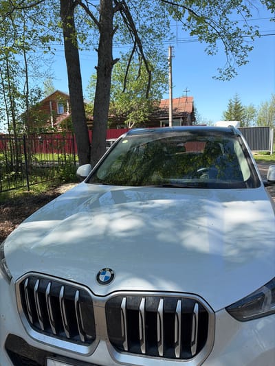 The image captures a close-up of the front of a white BMW car, parked outdoors. The BMW logo is visible on the front grille. The car's hood reflects the surrounding environment, including trees and a glimpse of a house with a red roof and red walls. A fence runs along the side of the house. In the background, clear blue sky peeks through the trees. Based on the available information, the location is Юг, Russia.