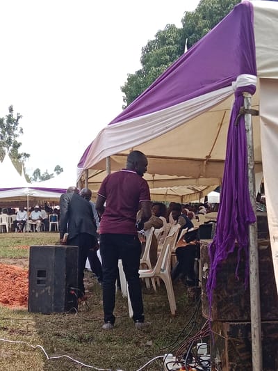 The image displays an outdoor event occurring on a grassy field in East Wanga ward, Kenya. Multiple event tents are present, including a prominent white and purple canopied structure in the foreground. A male subject wearing a maroon short-sleeved shirt and dark trousers stands with his back to the camera in the mid-foreground, facing towards a seated audience. To his immediate left, another male subject in a dark suit is partially visible, bending over an object near the ground. A large black speaker is positioned on the grass in the left foreground, connected by visible cables. Adjacent to the speaker is a quantity of reddish-brown, shredded material. In the right foreground, a vertical tent support pole wrapped with purple fabric stands next to a stack of dark equipment boxes. Tangled electrical wires and a power strip are visible on the ground at the base of this pole. Numerous individuals are seated on white plastic chairs arranged in rows under the tents, indicating a gathering. The background features additional tent structures, green foliage from trees, and a bright, open sky.