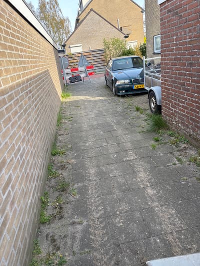 The scene depicts a narrow driveway or alleyway in Heemskerk, Netherlands. The alley is paved with grey interlocking blocks, and the bordering walls are constructed of red brick. Vegetation, including grass and weeds, grows sporadically along the edges of the paved area. A dark green BMW automobile with the license plate "SB-NJ-1" is partially visible, parked at the end of the alley. A trailer is parked behind the car. The surrounding buildings appear to be residential homes. The sky is partially visible in the upper left corner.