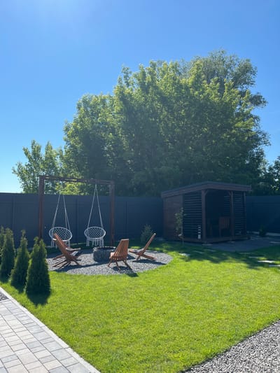 This is a wide, eye-level shot of a backyard in Sharovechka, Ukraine, under a clear blue sky. In the foreground, a paved walkway with alternating gray and beige square tiles runs diagonally from the bottom left. The walkway borders a meticulously manicured green lawn.

The middle ground of the image features a gravel area with a wooden swing set supporting two white macrame chairs. In front of the swings is a modern fire pit with a stone surround. Three wooden lounge chairs are arranged around the fire pit. To the right of the fire pit, a wooden gazebo with a dark roof and open slatted walls sits on a gravel foundation.

The background is dominated by a dark gray, solid fence that encloses the property. Beyond the fence, a dense canopy of lush green trees fills the upper portion of the image. Several small, conical evergreen trees are planted in a line along the left side of the lawn, near the fence. The lighting suggests it is a sunny day, with the sun casting shadows from the trees onto the lawn.