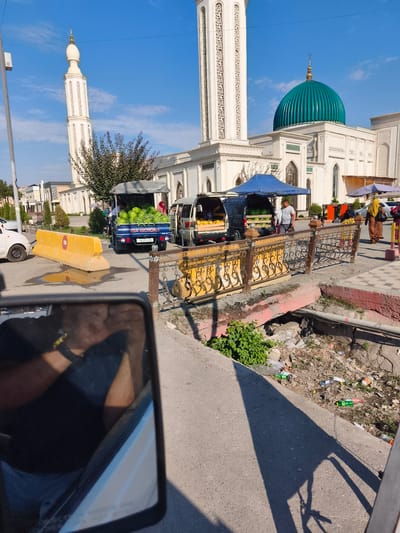 In the city of Buz, Uzbekistan, an outdoor market is set up in front of a large white mosque with a green dome. A minivan and a truck are parked side-by-side, laden with watermelons and other produce. People are seen browsing the market stalls and interacting with vendors. In the foreground, a car's side mirror reflects the interior, showing a driver and a passenger. A yellow concrete barrier is placed on the road, possibly for traffic control. The scene is illuminated by bright sunlight, casting shadows on the ground. The overall impression is one of a bustling market day in a vibrant urban setting.