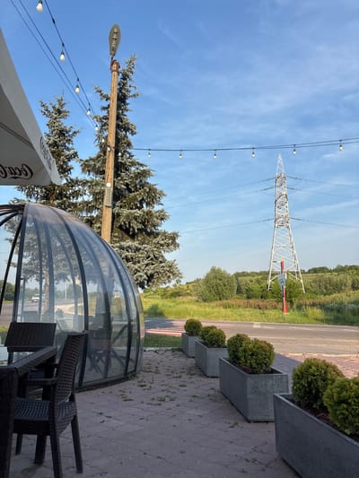 The image depicts an outdoor seating area adjacent to a glass-enclosed structure, possibly a cafe or restaurant. Several dark wicker chairs and tables are visible inside the glass structure, suggesting a place for patrons. Outside, concrete planters with neatly trimmed bushes line a paved walkway.

Dominating the background is a large, white metal power line pylon, a common sight in rural or semi-rural landscapes. A road runs parallel to the scene, bordered by grassy fields and lush green trees. The sky is a clear, bright blue, with a few wispy clouds, indicating fair weather. String lights hang across the sky, adding to the ambiance of the seating area. A large wooden pole with a street lamp and more string lights is also prominent in the foreground.

The overall impression is that of a pleasant outdoor dining or relaxation spot, set against a backdrop of power infrastructure and natural scenery. The time of day appears to be late afternoon or early evening, given the angle of the sunlight casting subtle shadows. No people are visible in the scene. Text is visible on a white umbrella in the lower left corner, appearing to be a Coca-Cola logo.