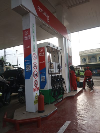 The image depicts a Pertamax gas station in Tocina, Indonesia. In the foreground, a red fire extinguisher stands upright on a raised concrete platform. A gas pump with multiple hoses is visible to the left. Two individuals in red uniforms, presumably gas station attendants, are present. One attendant, wearing a red cap and uniform, is standing next to a motorcycle on the right side of the frame. The other attendant, also in a red uniform, is facing away from the camera and leaning against the gas pump. Several motorcycles are parked at the station, including one in the left foreground with a license plate reading "DW 4663 BD". A building with a light-colored facade and arched windows is visible in the background. The sky is bright and overcast. Several warning signs are posted on the gas pump pillars, including ones prohibiting smoking, photography, and cell phone use. The ground is paved with a reddish-brown material and shows signs of wetness, reflecting the overhead lighting.