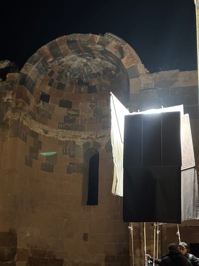 A low-angle shot depicts a section of a stone structure at night. The structure appears to be a tower or dome, constructed from alternating bands of light and dark stone. To the right, a large, rectangular black screen is suspended, illuminated by a bright light source positioned behind it. Part of a lighting rig is visible, with a man in a dark jacket and light-colored baseball cap kneeling near the rig. The background is dark, suggesting it is night. The image may be of a section of a church in the city of Anipemza, Armenia, as the architectural style resembles structures found there.