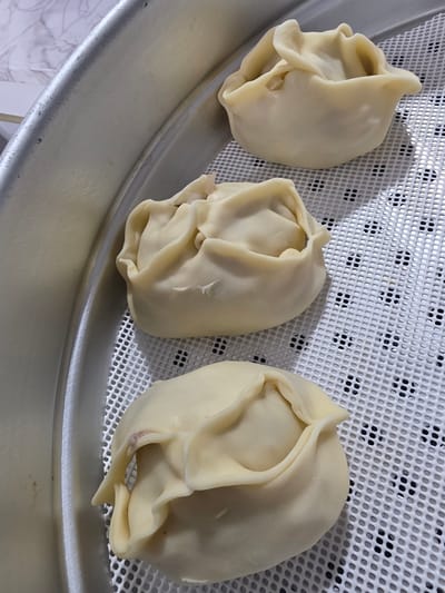 A close-up, top-down view shows three uncooked manti dumplings arranged in a steamer basket. The dumplings are pale yellow and have a ruffled, folded appearance. They sit on a white, perforated plastic mat, which rests inside a silver steamer tray. The background is a blurred marble-patterned surface. The image was taken indoors, likely in a kitchen, and does not provide any information about the city of Eumseong-eup, South Korea.