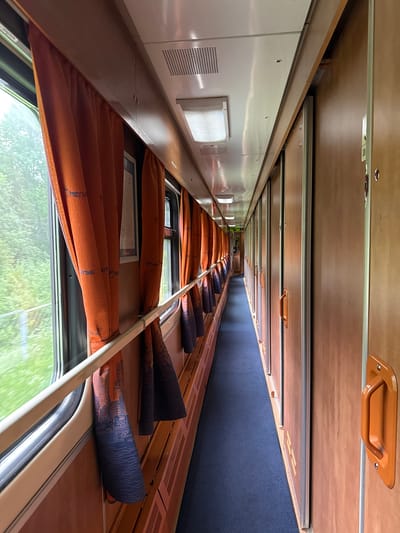 The image displays the interior of a train carriage, likely a long-distance or sleeper coach, viewed along its length in a long, narrow perspective. The setting is a train traveling through a green, natural landscape, possibly near Krasnoborskoye, Russia, given the visible Cyrillic text.
On the left side of the carriage, a row of windows is visible. Most windows are covered by pleated, orange-brown curtains with blue geometric patterns, some partially rolled up, others fully extended. Reflections of blurred green foliage, indicative of movement, can be seen through the windows. A narrow wooden or laminate ledge runs along the base of the windows. A white metal bar extends horizontally across the window area. Visible text on the curtains includes "МЕГАТРАНС", "МЕТАЛЛИС", and "ЭТАЖ".
On the right side, there is a long, dark blue upholstered wall or partition, possibly serving as backrests or headboards for lower bunks. Above this, a light brown wooden panel runs the length of the carriage, equipped with small orange handles, suggesting the presence of upper bunks or overhead storage.
The ceiling consists of white panels featuring rectangular fluorescent light fixtures and perforated grilles, likely for air conditioning or ventilation. A small panel near the foreground shows the numbers "23". The floor is not fully visible but appears to be a dark surface, possibly a blue carpet, towards the far end of the carriage.
The lighting suggests it is daytime, with natural light entering through the windows complemented by interior artificial lighting. No people are visible, and the scene conveys a sense of quiet travel.