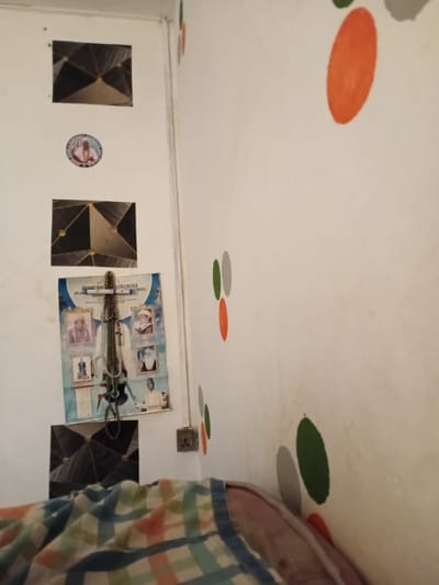 The image depicts an interior scene, possibly a bedroom, with white walls serving as the backdrop. On the left wall, there are several decorative elements: two framed geometric patterns, one circular sticker with an image of a person, and a poster-like item with multiple portraits and what appear to be Islamic inscriptions, adorned with several strands of prayer beads. To the right of the poster, an electrical outlet is visible. The wall on the right side of the image features a pattern of colorful oval shapes in red, green, and gray. In the foreground, a portion of a blanket or comforter with a plaid pattern of blue, white, orange, and green is visible, resting on what seems to be a bed. The overall impression is of a modestly decorated living space. Without further information or visual cues, it is not possible to definitively place this scene in Ipota, Nigeria.