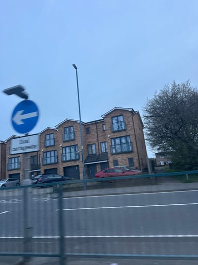 The scene depicts a residential area adjacent to a dual carriageway under an overcast sky, viewed from a moving vehicle. The location is Gillingham, United Kingdom, specifically alongside a segment of Maidstone Road (A229) in proximity to "The Cottageway" development, as indicated by partially legible roadside signage.
The environment features a multi-lane roadway in the foreground, marked by white lines, separated from a raised embankment by a metal guardrail. A tall, slender grey street lamp stands prominently in the mid-ground. The primary subjects are a row of at least four modern, three-story brick townhouses. Each unit exhibits dark-framed windows, many equipped with dark metal Juliet balconies. The ground floors feature dark garage doors and entryways. Roofs are dark-tiled with distinct gables.
A blurred blue circular road sign with a white left-pointing arrow is visible on the far left. Below this, a white rectangular sign partially displays the text "Dual Carriageway" and "That Cottageway". Four parked vehicles are visible on the raised area near the houses: a silver hatchback (partially obscured, left), a dark-colored sedan (left-center), a red sedan (center-right), and a second silver vehicle (partially obscured, right). A large, bare deciduous tree is positioned on the right, behind the residential structures, with some underlying green shrubbery. No individuals are present, and no active interactions are depicted.
