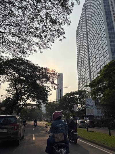 The image captures a street scene in Kelapa Dua, Indonesia, on a bright, hazy day. Tall, modern buildings with many windows dominate the right side of the frame, while silhouetted trees with dense foliage frame the top and left. The sun is visible in the sky, partially obscured by tree branches, casting a warm glow.

In the foreground, a person is riding a motorcycle, wearing a blue denim jacket and a pink helmet. A black car is positioned to the left, and another motorcycle with a rider wearing a helmet is in front of the depicted motorcycle. In the middle ground, several other vehicles, including cars and motorcycles, are visible on the road, moving in the same direction. Pedestrians can be seen further down the street.

On the right side of the road, there are commercial establishments with signs, one of which advertises "Gramedia Back to School" with a 50% discount. Green grass and trees line the median and the side of the road. The overall impression is of a busy urban environment during daylight hours, with a mix of natural elements and modern infrastructure.