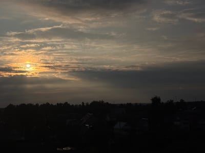This media file captures a twilight scene over a rural Belarusian landscape, likely within the Pratasewicki rural council. The sun, low on the horizon and partially obscured by clouds, casts a warm, golden light that illuminates the sky with streaks of yellow, orange, and white. The clouds themselves are varied, with some appearing thin and wispy, catching the light, while others are darker and more dense, creating a dramatic contrast.
Below the sky, a dense line of dark trees forms the treeline of a village. The village itself is rendered in deep shadow, with the silhouettes of numerous houses and buildings visible. Some of the roofs show faint glints of light, suggesting they might be made of reflective materials or are catching ambient light. The overall impression is one of a quiet, dusk setting.
The time of day is clearly either sunrise or sunset, given the low angle of the sun and the warm, diffused light. The weather appears to be partly cloudy, with the clouds playing a significant role in shaping the light and mood of the scene. The location cues, such as the style of the houses and the general landscape, are consistent with a rural area in Eastern Europe, and specifically Belarus. There is no visible text in the image. The mood is peaceful and serene, with a touch of melancholy often associated with twilight.