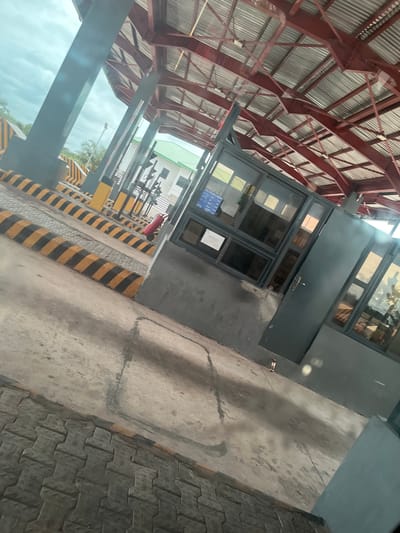 A street-level, angled outdoor medium shot captures a view of a toll plaza on a cloudy day. The foreground features a grey concrete surface with interlocked brick paving on the left and yellow and black striped concrete barriers. In the middle ground, a grey booth with large windows is visible, along with a dark grey door. A red fire extinguisher on wheels stands to the left of the booth. In the background, the toll plaza's corrugated metal roof with red support beams is prominent. Beyond the plaza, a white building with a green roof and some trees can be seen. The scene suggests a functional toll collection point in an urban or semi-urban environment.