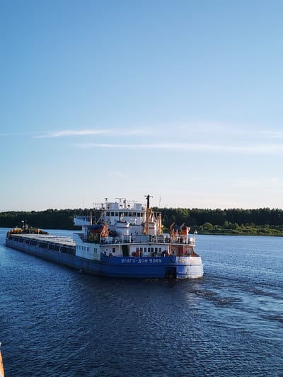 A wide blue river flows with gentle waves under a clear, light blue sky with a few wispy white clouds. The sun is shining, suggesting it's daytime. On the right side of the river, a dense, dark green forest lines the bank.
Dominating the foreground and midground is a long, dark blue barge being pushed by a white tugboat. The barge is filled with cargo, covered by a large blue tarp. The tugboat is white with blue accents and has several decks and railings. On the side of the tugboat, the text "ВОЛГО-ДОН 5069" is visible in white lettering.
Several people are visible on the decks of the tugboat, looking out at the surroundings. Some life preservers in orange and blue are mounted on the boat.
To the left of the main barge, a smaller, yellow vessel is also visible on the water, possibly another part of the same convoy or a separate boat.
The overall scene depicts a tranquil river journey, likely in Russia given the Cyrillic text on the boat and the mention of Sud'bitsy. The clear sky and calm water suggest pleasant weather. The activity is the movement of the barges and tugboat along the river.